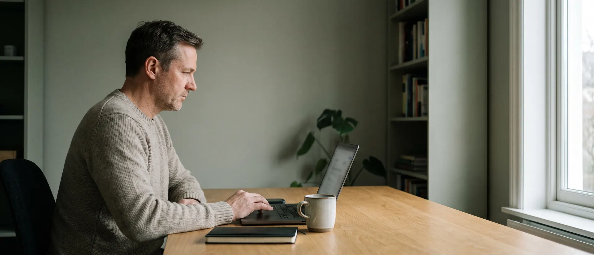 An engineer working side-on at a laptop in a plain home office, lit by grey daylight from a window.